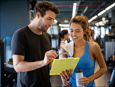 A man and woman in a gym, both holding a tablet while exercising at a Wellbridge fitness center